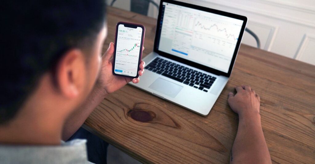 Individual reviewing stock market trends using a laptop and smartphone at a wooden table.